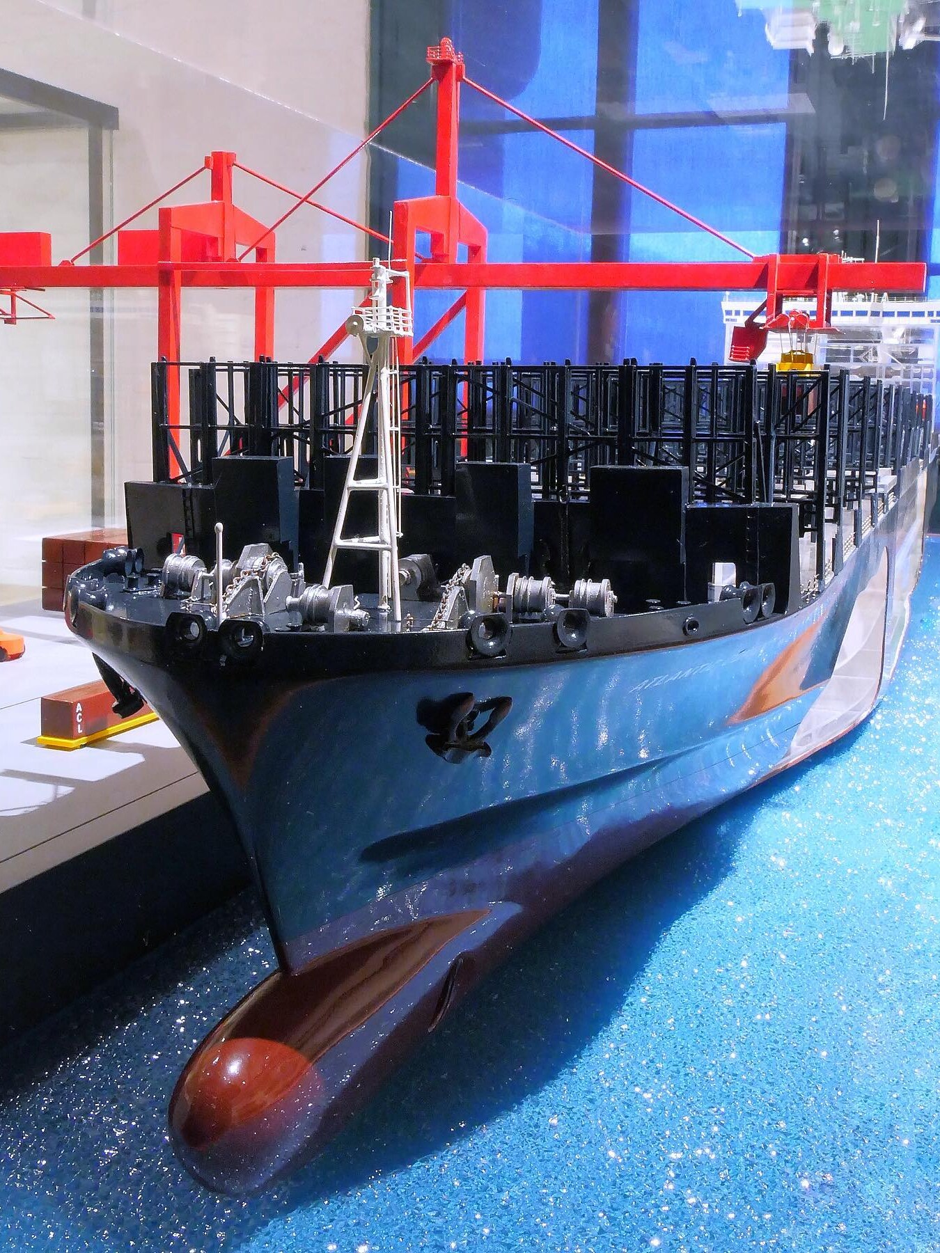 Modell eines Containerschiffs am Kai mit sichtbarem Bug, Ankerwinden und roter Hafenkran-Konstruktion im Hintergrund