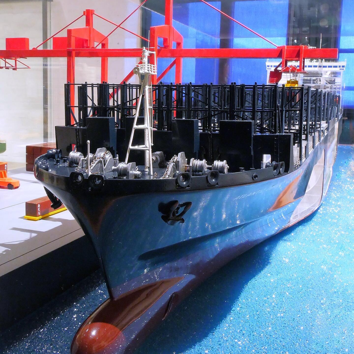 Modell eines Containerschiffs am Kai mit sichtbarem Bug, Ankerwinden und roter Hafenkran-Konstruktion im Hintergrund