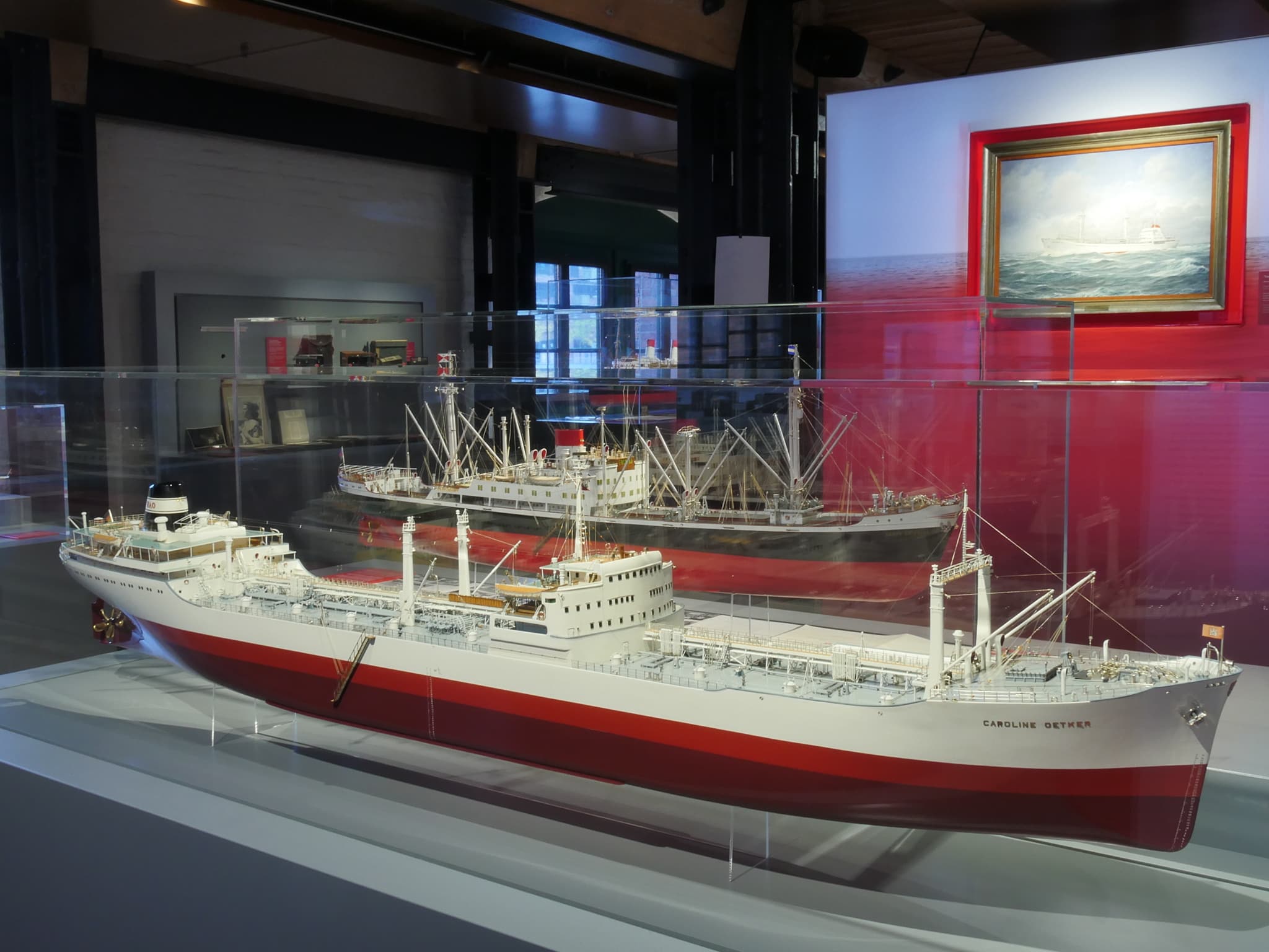 Zwei detaillierte Schiffsmodelle mit rot-weißen Rümpfen in einer Glasvitrine eines Museums, im Hintergrund ein weiteres Modell und ein maritimes Gemälde.