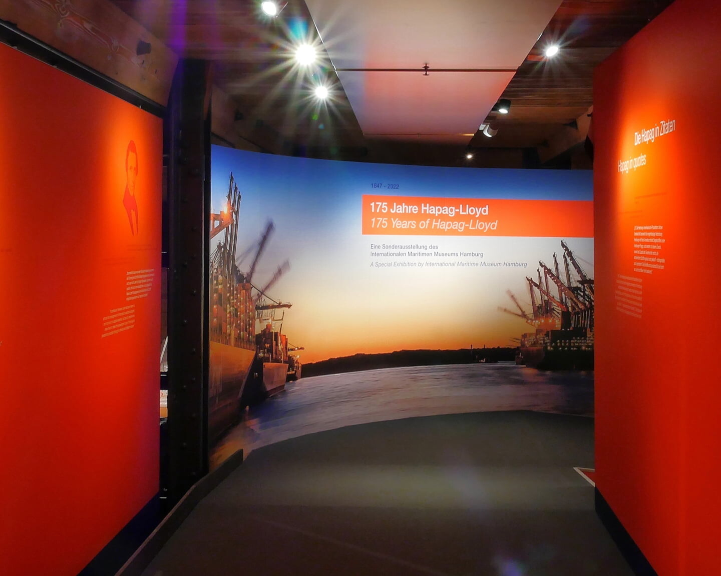 Ausstellungsflur mit orangen Wänden und Portraits, am Ende Tafel '175 Jahre Hapag‑Lloyd' vor Containerschiffen und Hafenkränen im Abendlicht.