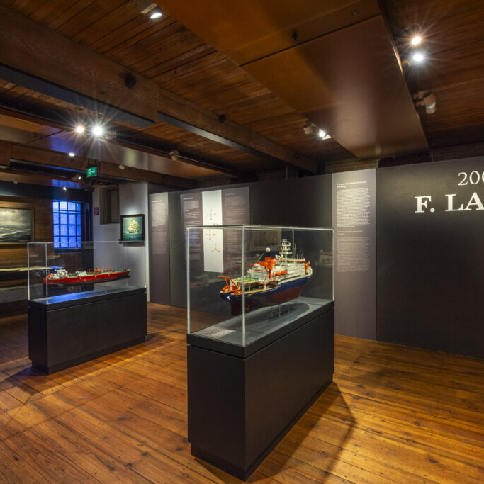 Hölzerner Museumsraum mit sichtbaren Deckenbalken, zwei Schiffsmodelle in Glasvitrinen auf dunklen Sockeln, warmes Ausstellungslicht, maritime Bilder an den Wänden und die große Wandbeschriftung