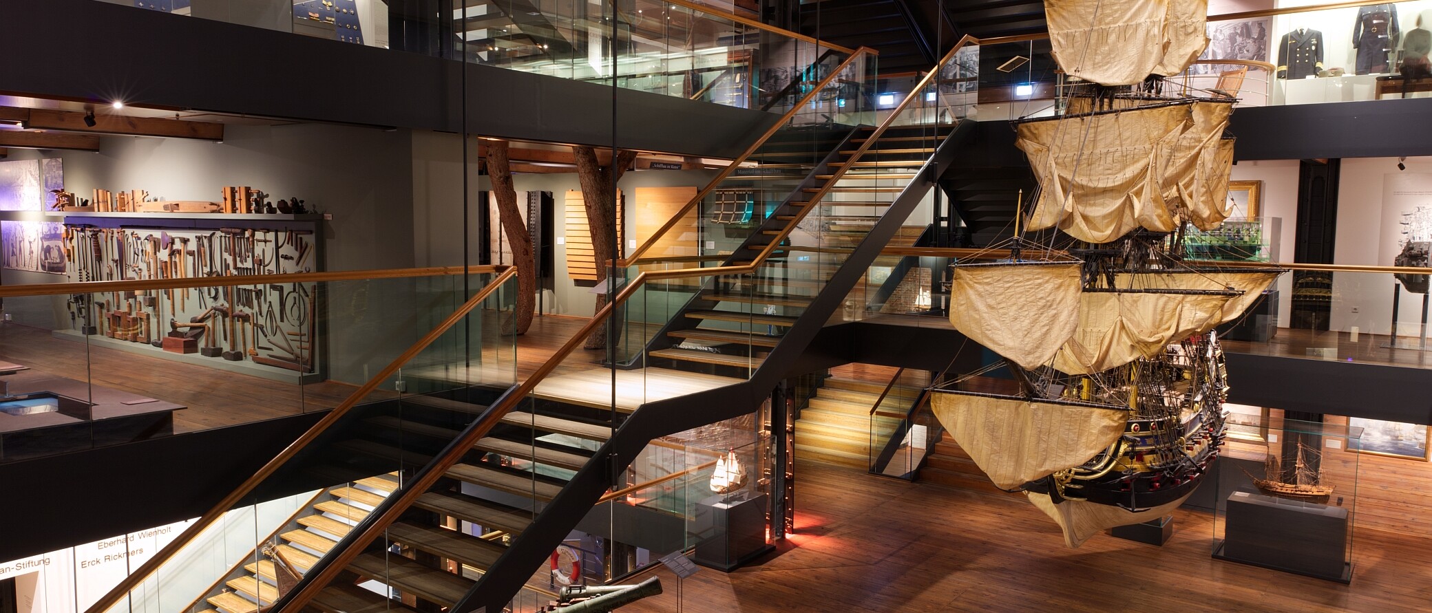 Gro&szlig;es h&auml;ngendes Modellsegelschiff mit beigen Segeln in einem Museum mit Glas-Holz-Treppen.