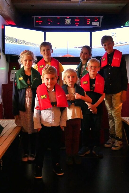 Sieben Kinder mit roten und grünen Rettungswesten stehen in einem Schiffssimulator vor drei Bildschirmen, umgeben von Holzbänken und maritimen Wandbildern.