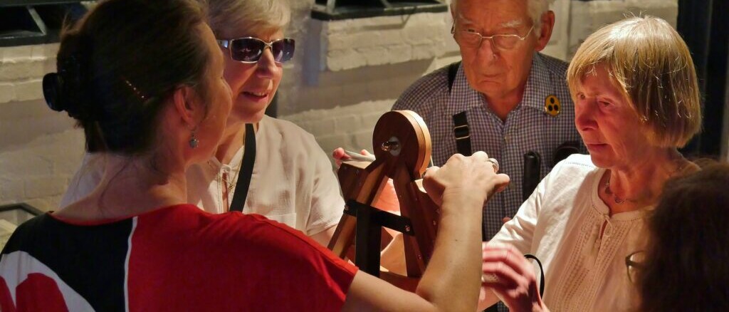 Vier ältere Erwachsene umringen ein hölzernes mechanisches Modell mit Scheibe und Kurbel in einem Museumsraum.