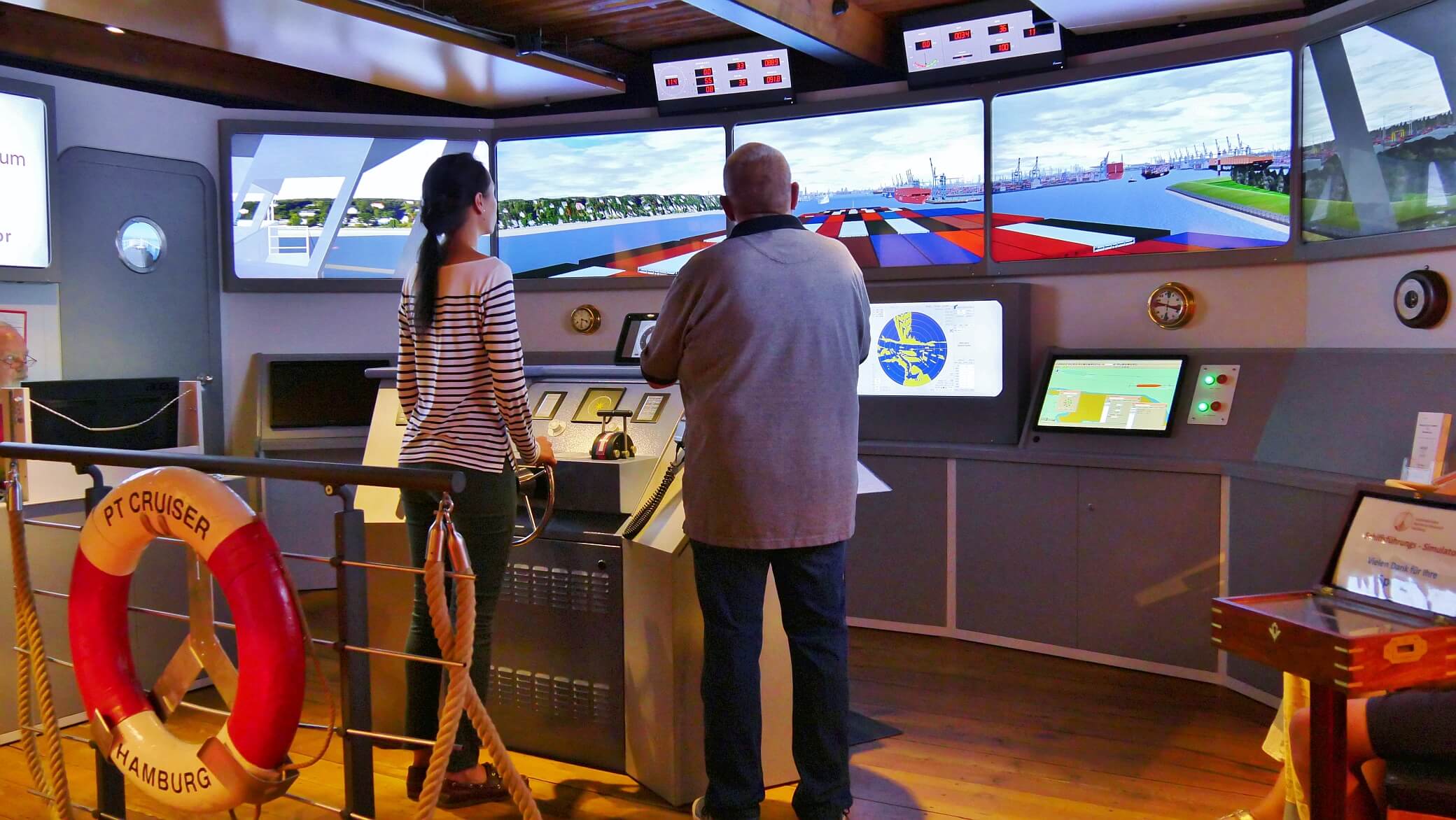 Zwei Personen an Br&uuml;ckensimulator vor Panorama-Monitoren mit Containerschiffansicht und Rettungsring mit Hamburg-Schriftzug.