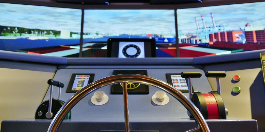 Blick &uuml;ber ein poliertes Metallsteuerrad auf die Br&uuml;cke eines Schiffs-Simulators mit Schubhebeln, Kn&ouml;pfen und mehreren Bildschirmen, die einen Hafen mit Containern zeigen.