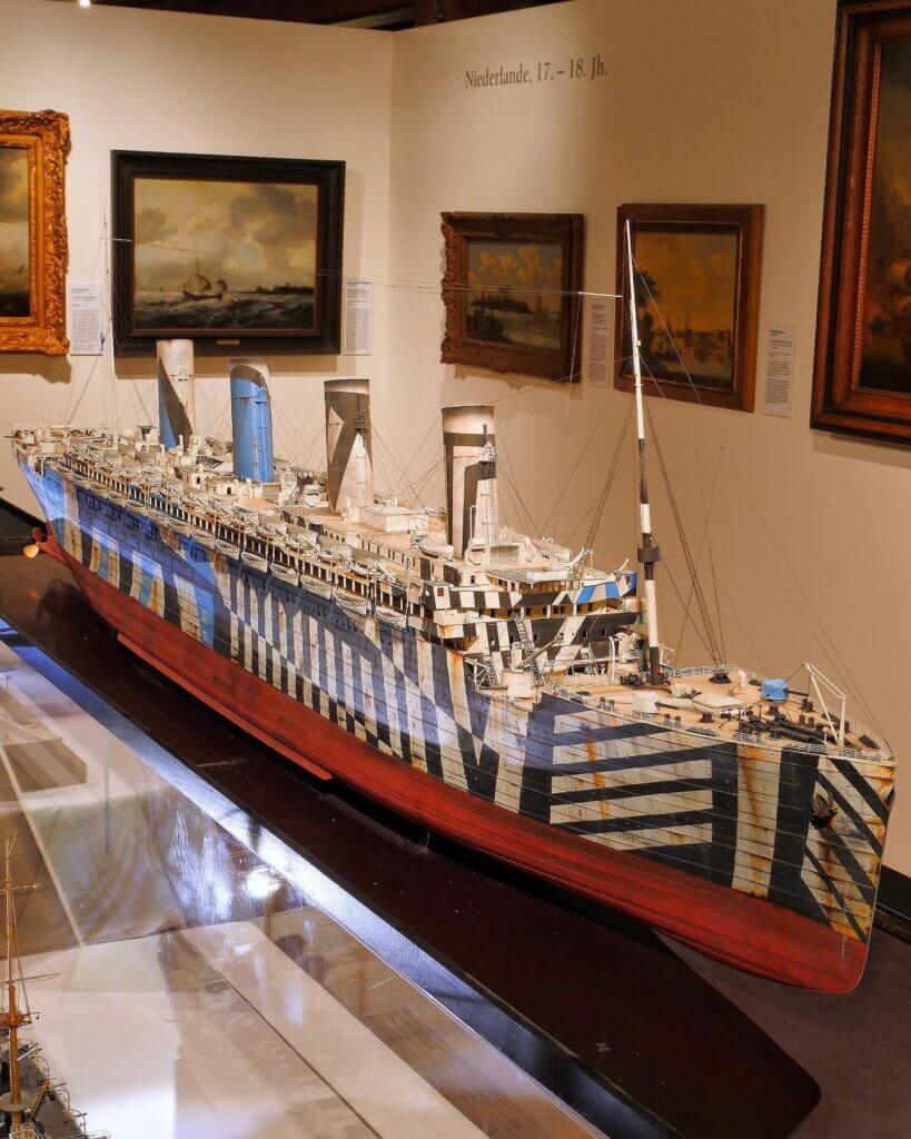 Großes Schiffsmodell in einem Museum mit auffälliger Dazzle-Tarnung aus schwarzen und weißen geometrischen Streifen, rotem Rumpf und drei Schornsteinen.