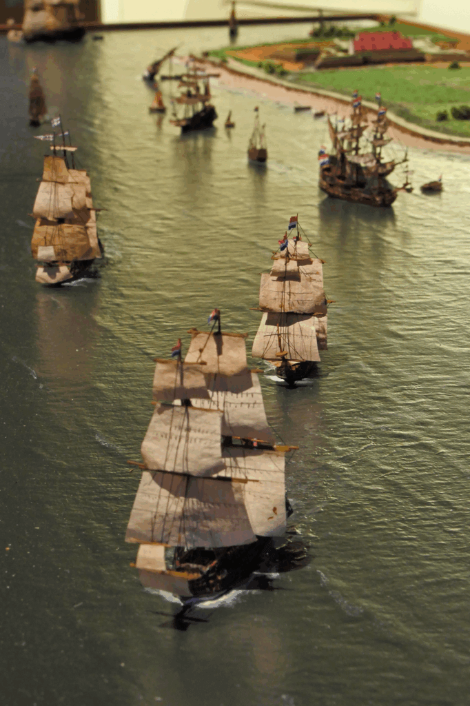 Diorama mit mehreren historischen Dreimast-Segelschiffen in Formation auf ruhigem Wasser, mit Booten und einer grünen Küstenlandschaft im Hintergrund.