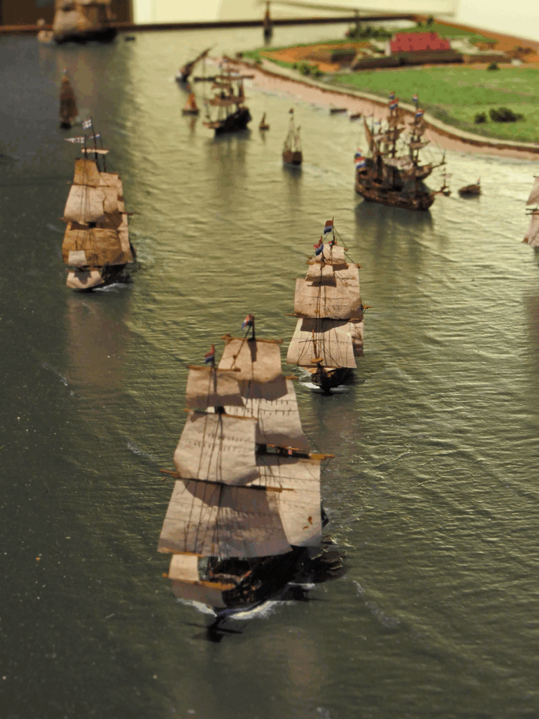 Diorama mit mehreren historischen Dreimast-Segelschiffen in Formation auf ruhigem Wasser, mit Booten und einer grünen Küstenlandschaft im Hintergrund.