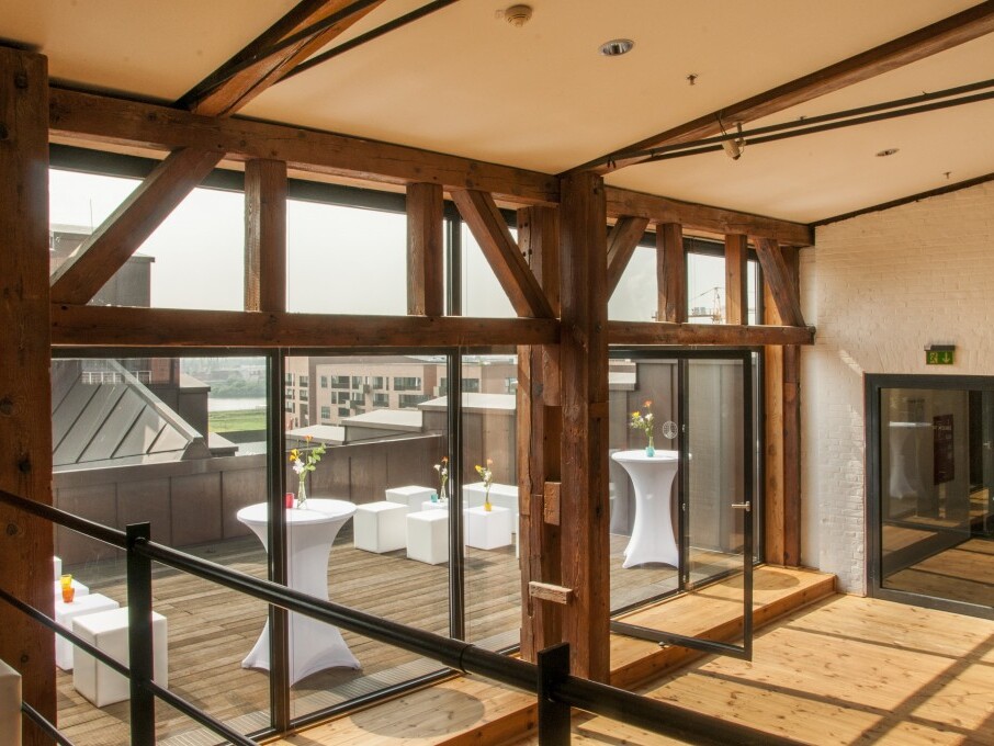 Heller Raum mit Holzträgern und großer Glasfront, Blick auf Dachterrasse mit Stehtischen und weißen Sitzwürfeln.