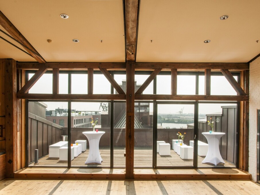 Großer Raum mit Holzbalken und bodentiefen Fenstern zu einer Dachterrasse mit weißen Stehtischen, Sitzwürfeln und Hafenblick.