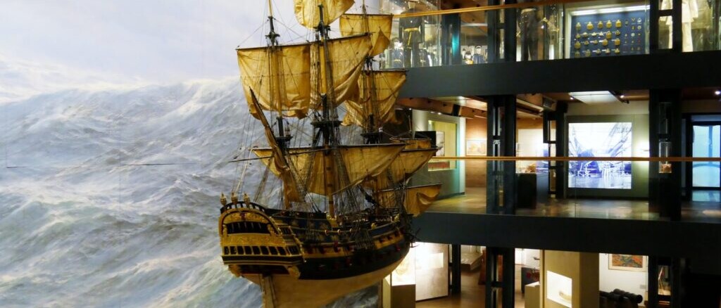 Hängendes Modell eines Segelschiffs mit geblähten Segeln vor bemaltem Sturmmeer in einem mehrstöckigen Museum.