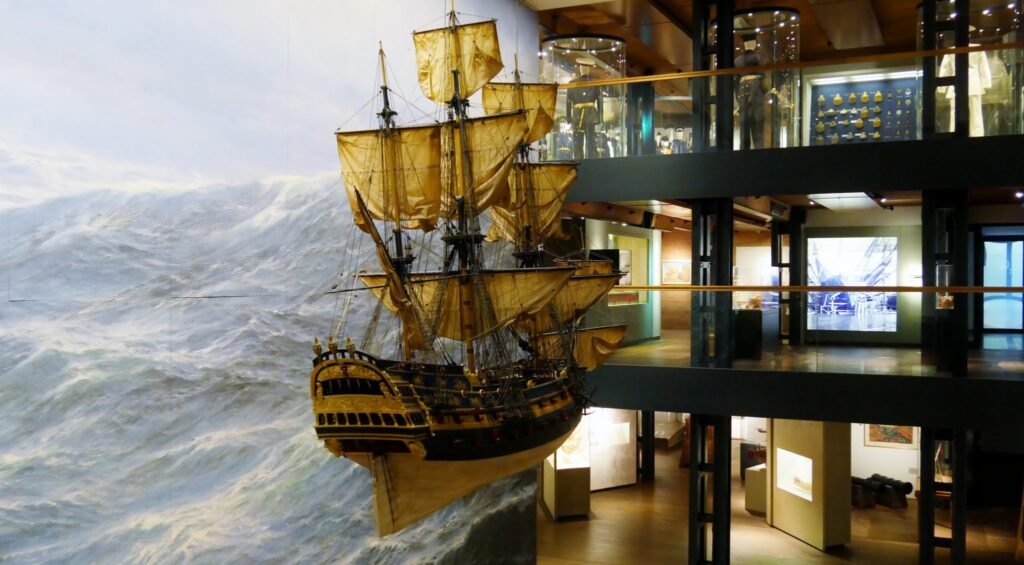 Hängendes Modell eines Segelschiffs mit geblähten Segeln vor bemaltem Sturmmeer in einem mehrstöckigen Museum.