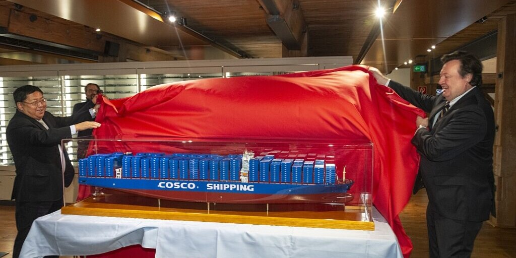 Zwei Männer lüften ein großes rotes Tuch und enthüllen auf einem weißen Tisch ein im Glaskasten ausgestelltes Modell-Containerfrachtschiff mit blauen Containern und der Aufschrift COSCO SHIPPING in einem holzverkleideten Innenraum.