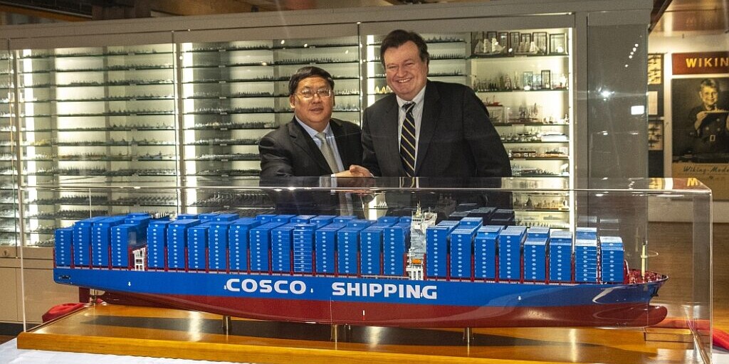 Zwei Männer in Anzügen schütteln lächelnd die Hand hinter einer großen blau-roten Modellvitrine des Containerschiffs mit der Aufschrift COSCO SHIPPING; im Hintergrund sind beleuchtete Vitrinen mit zahlreichen Schiffsmodellen zu sehen.