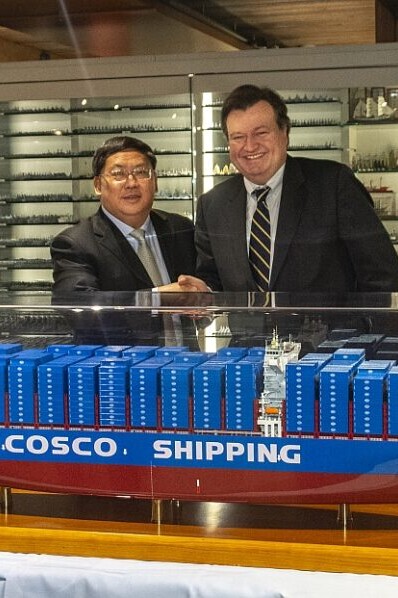 Zwei Männer in Anzügen schütteln lächelnd die Hand hinter einer großen blau-roten Modellvitrine des Containerschiffs mit der Aufschrift COSCO SHIPPING; im Hintergrund sind beleuchtete Vitrinen mit zahlreichen Schiffsmodellen zu sehen.