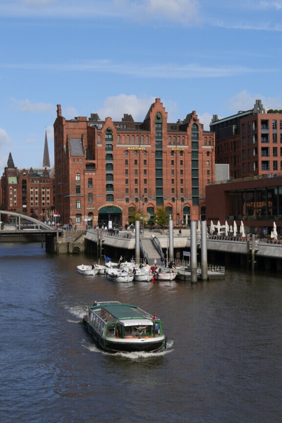 Grünes Passagierschiff fährt in einem Hafenkanal unter blauem Himmel; im Hintergrund ein großes rotes Backsteingebäude mit Giebeln, Schriftzug „Maritimes Museum“, Anleger und Yachten.