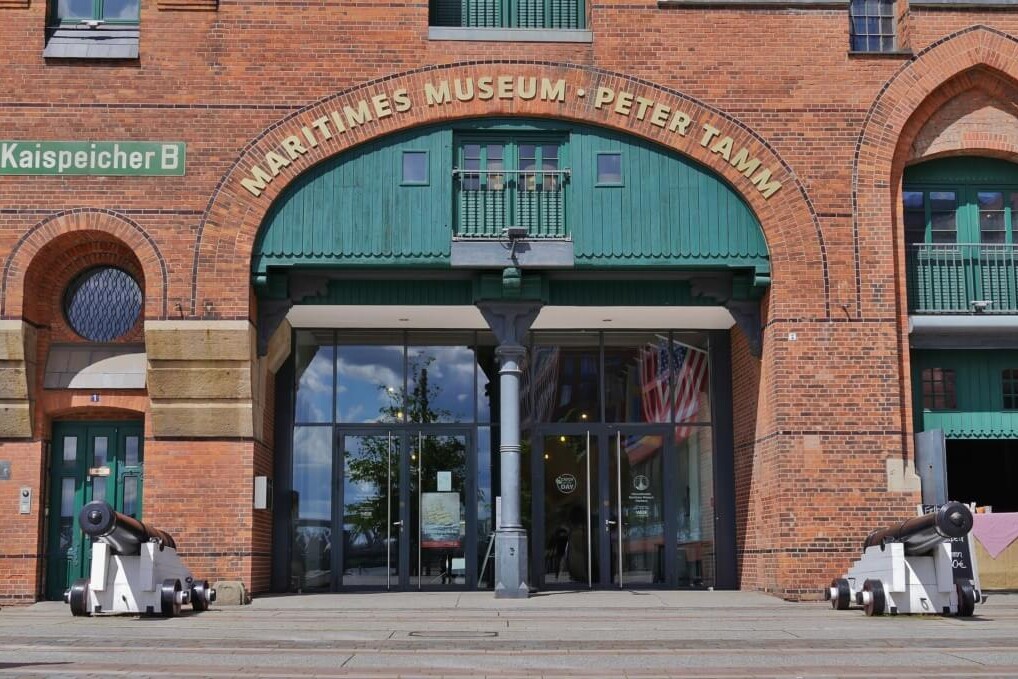 Eingang des Maritimen Museums Peter Tamm in rotem Backsteingebäude mit grünem Bogenportal, zwei historischen Kanonen links und rechts und Glas-Eingangstüren.