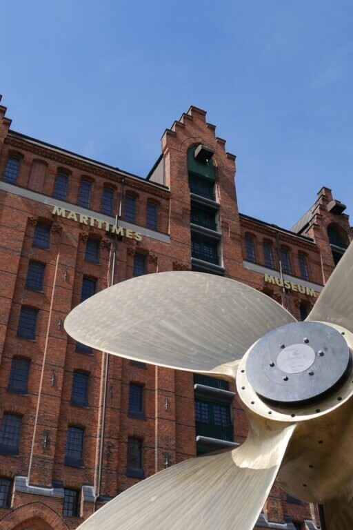 Großer bronzefarbener Schiffsschraubenpropeller im Vordergrund vor einem roten Backsteingebäude des Maritimen Museums unter klarem blauem Himmel.