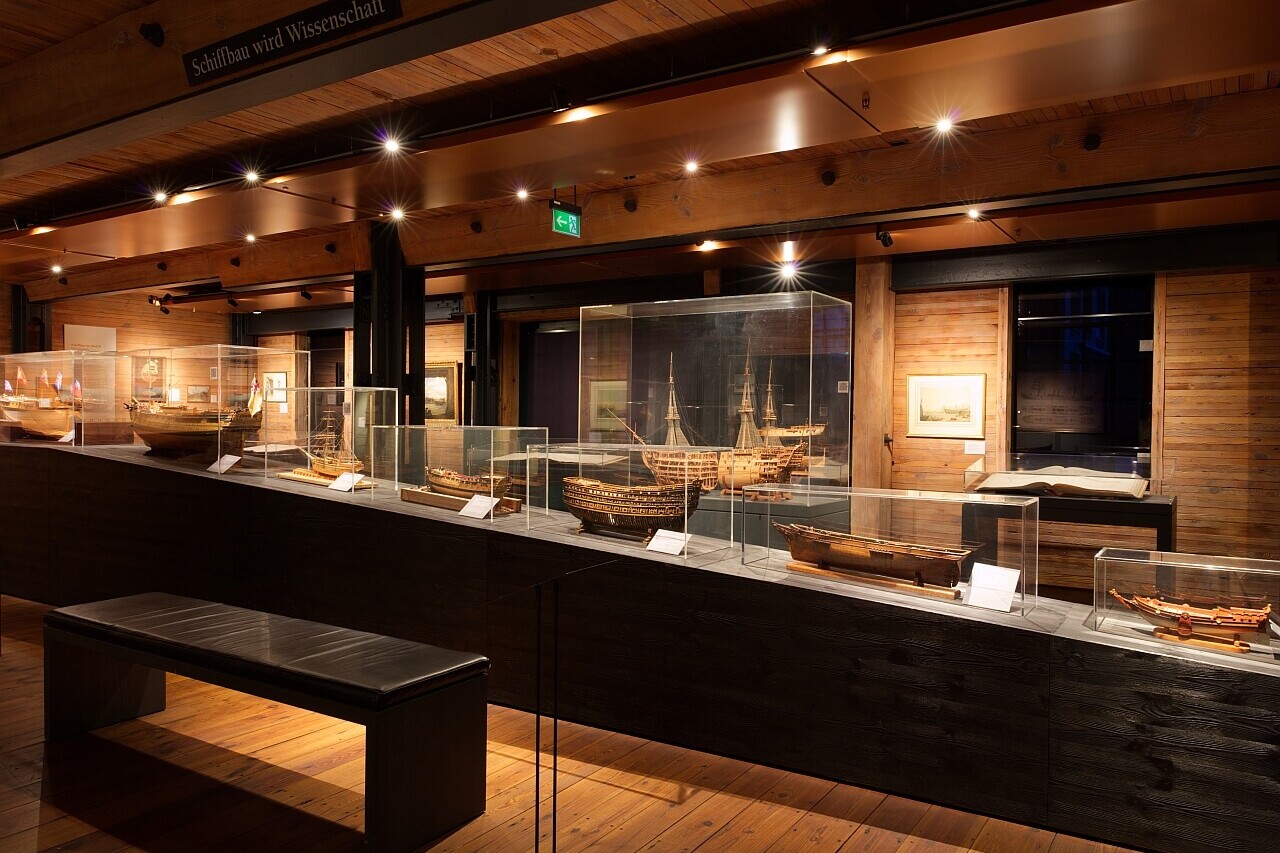 Dunkles Holzmuseum mit warmen Scheinwerfern, Glasvitrinen auf schwarzem Podest zeigen historische Schiffsmodelle; Vordergrund Sitzbank, Wände mit Bildern.