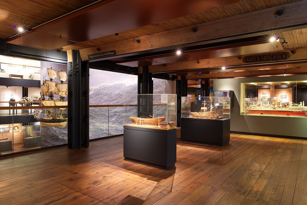 Museumshalle mit Holzfußboden und Balkendecke, Glasvitrinen mit Schiffsmodellen, großes Meereswandbild im Hintergrund und warme, gedämpfte Beleuchtung.
