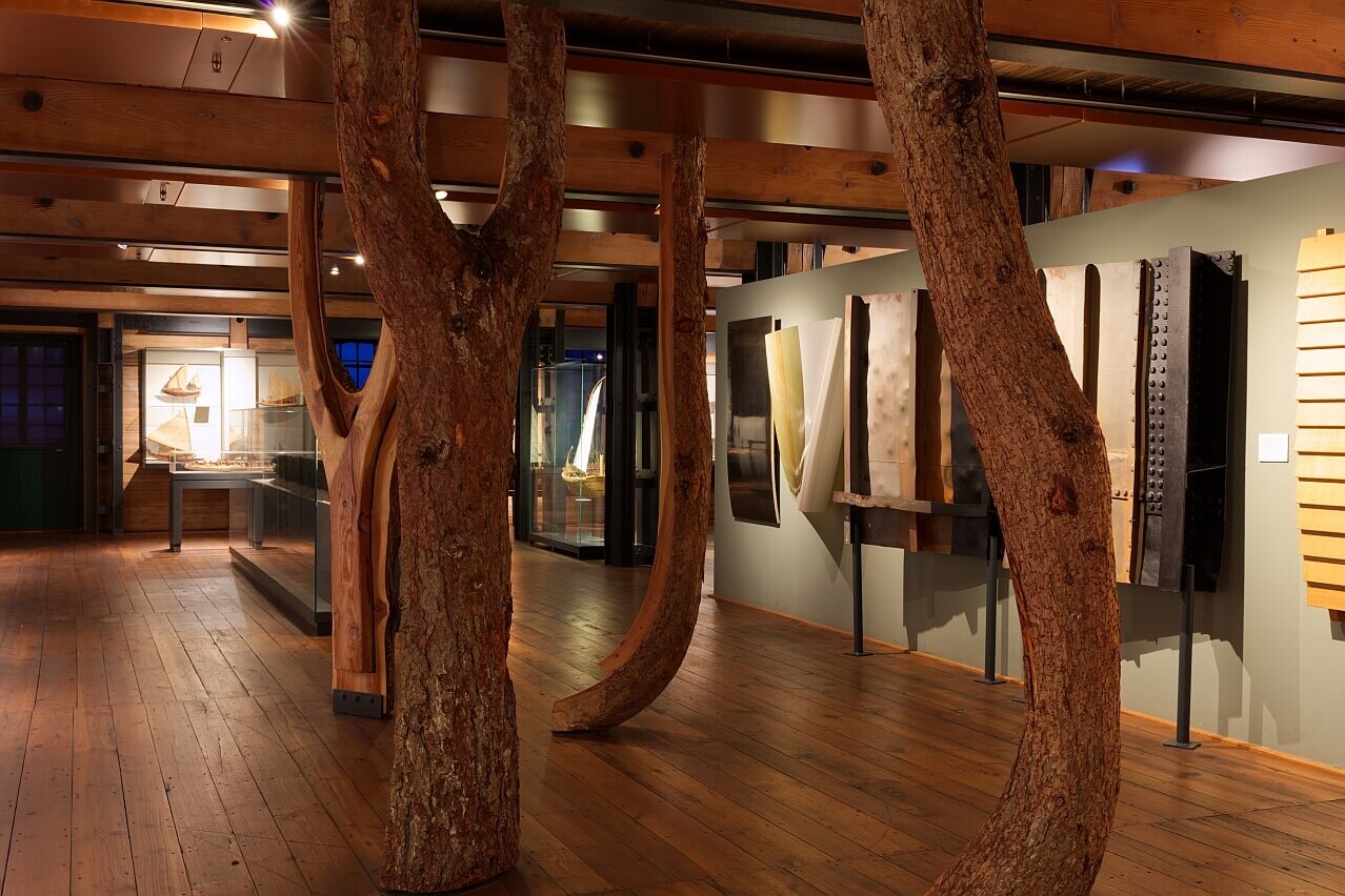 Museumsausstellung in warmem Licht mit massiven Baumstämmen als Säulen zwischen Holzbohlenboden, Vitrinen mit Schiffsmodellen und hölzernen Ausstellungstafeln.
