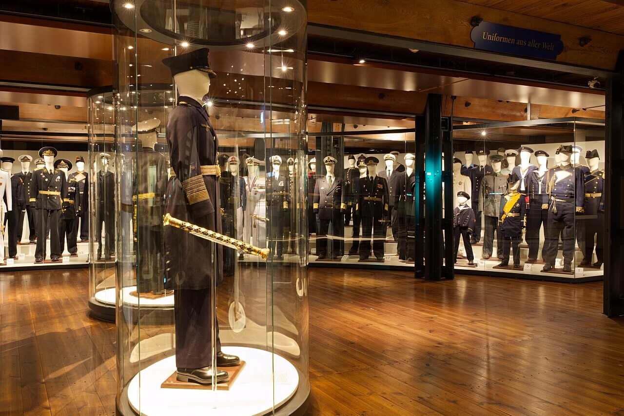 Museumsraum mit Mannequins in Milit&auml;r- und Marineuniformen; im Vordergrund ein Offiziersmannequin in rundem Glaskolben mit goldverziertem Degen.
