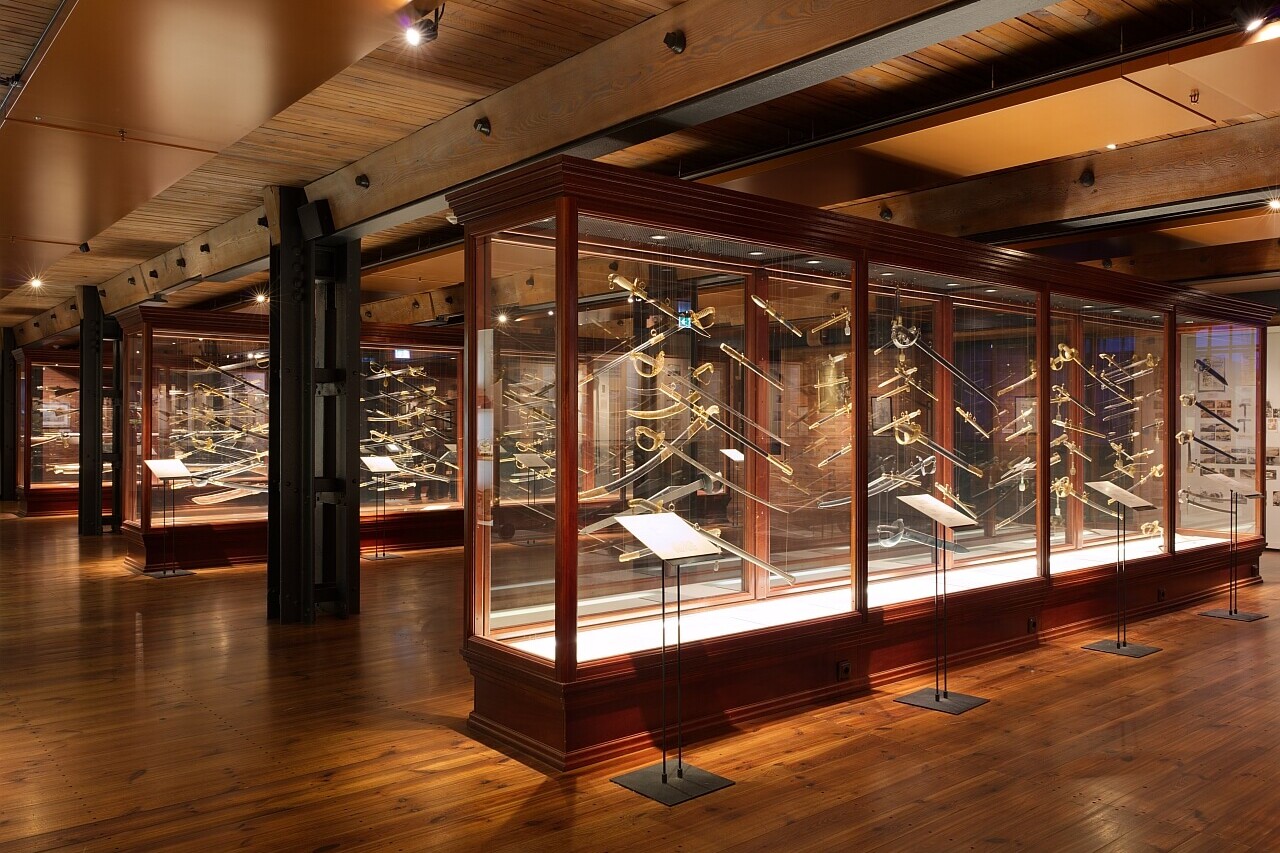 Weitl&auml;ufige Museumshalle mit warmem Licht und Holzboden, gro&szlig;e Glasvitrinen, darin zahlreiche historische S&auml;bel, Degen und Paradewaffen mit goldenen Griffen.