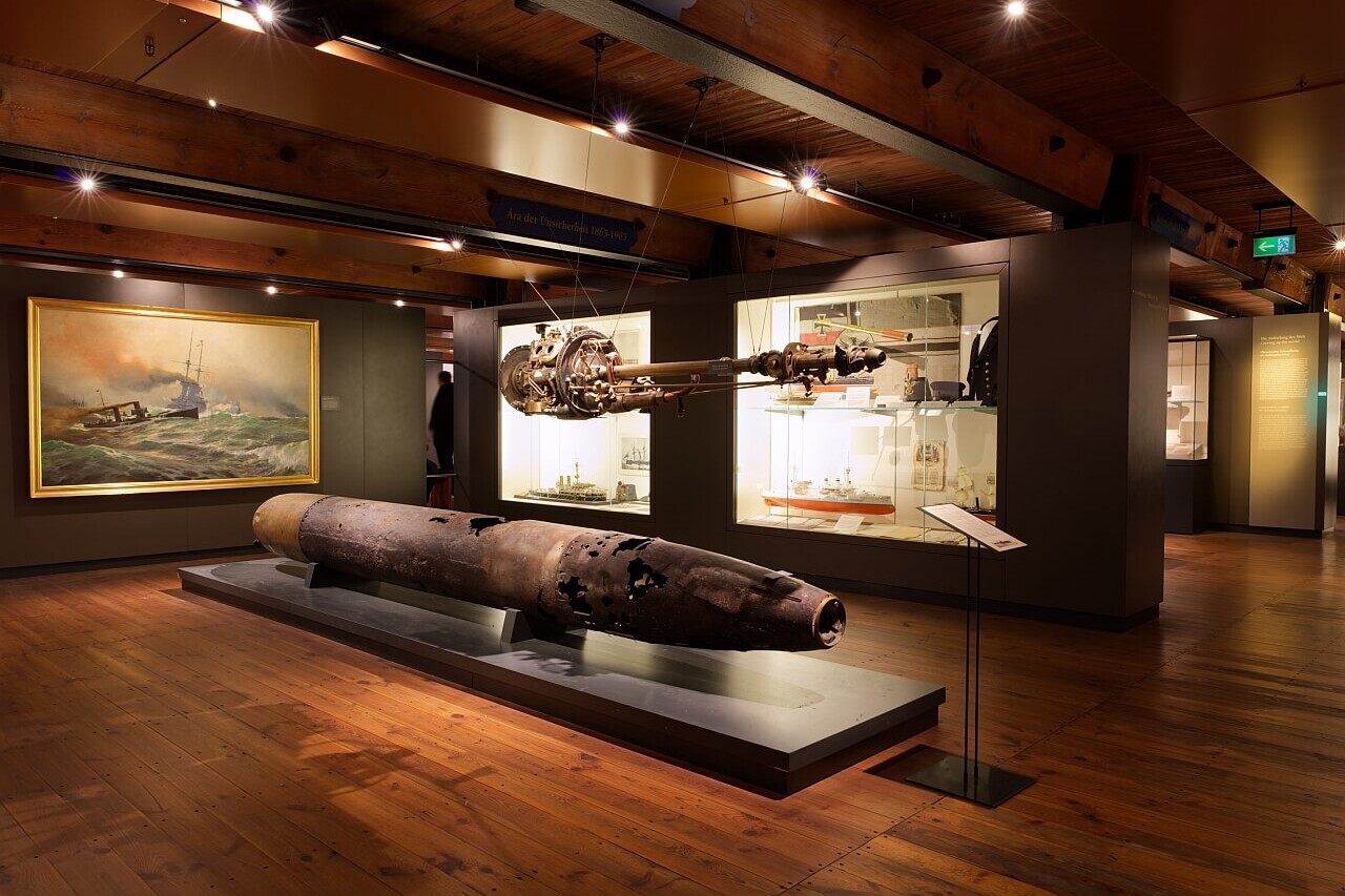 Museumsraum mit verrostetem Torpedo auf Podest, h&auml;ngendem Antrieb, Vitrinen mit Schiffsmodellen und Seeschlachtgem&auml;lde an der Wand.