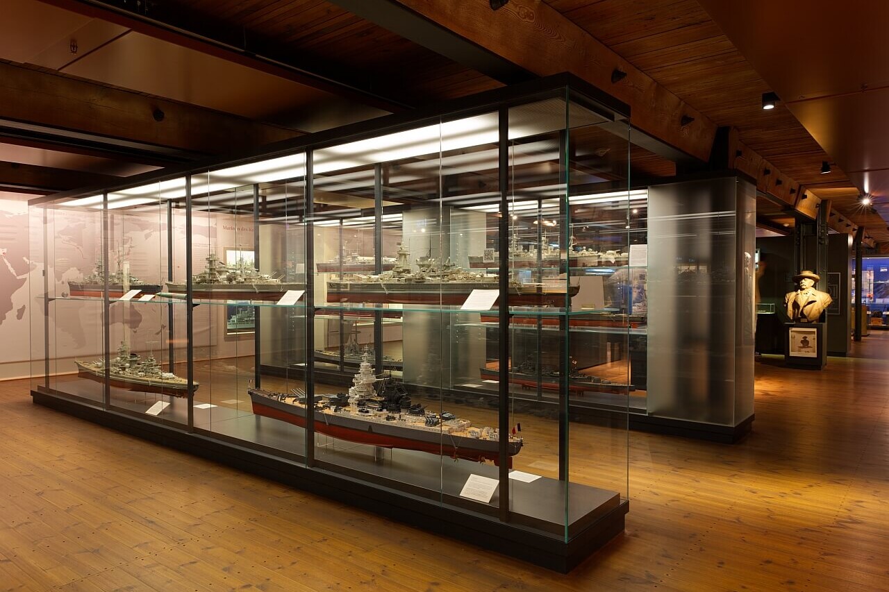 Museumsausstellung mit beleuchteten Glasvitrinen, in denen Schiffsmodelle stehen; warmer Holzboden und Deckenbalken, B&uuml;ste im Hintergrund.