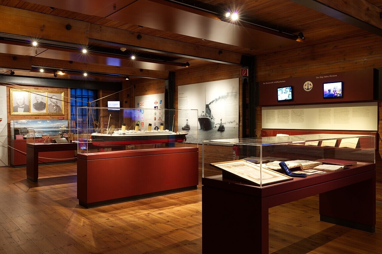 Warm beleuchteter Ausstellungsraum mit sichtbaren Holzbalken und Boden, Vitrinen mit Schiffsmodellen, Dokumenten, historischen Fotografien und Infotafeln.