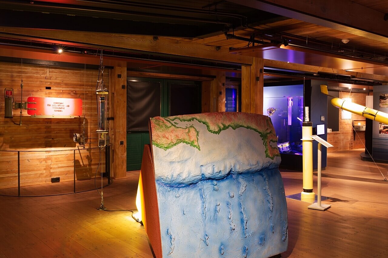 Warm beleuchteter Museumsraum mit Holzdecken, im Vordergrund ein großformatiges blau-grünes Relief des Meeresbodens und der Küstenlinie, umgeben von Messinstrumenten.
