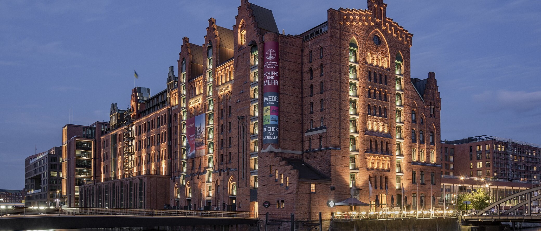 Beleuchtetes historisches rotbraunes Backsteingeb&auml;ude im Lagerhausstil am Wasser, mit Lichterspiegelung auf der Oberfl&auml;che