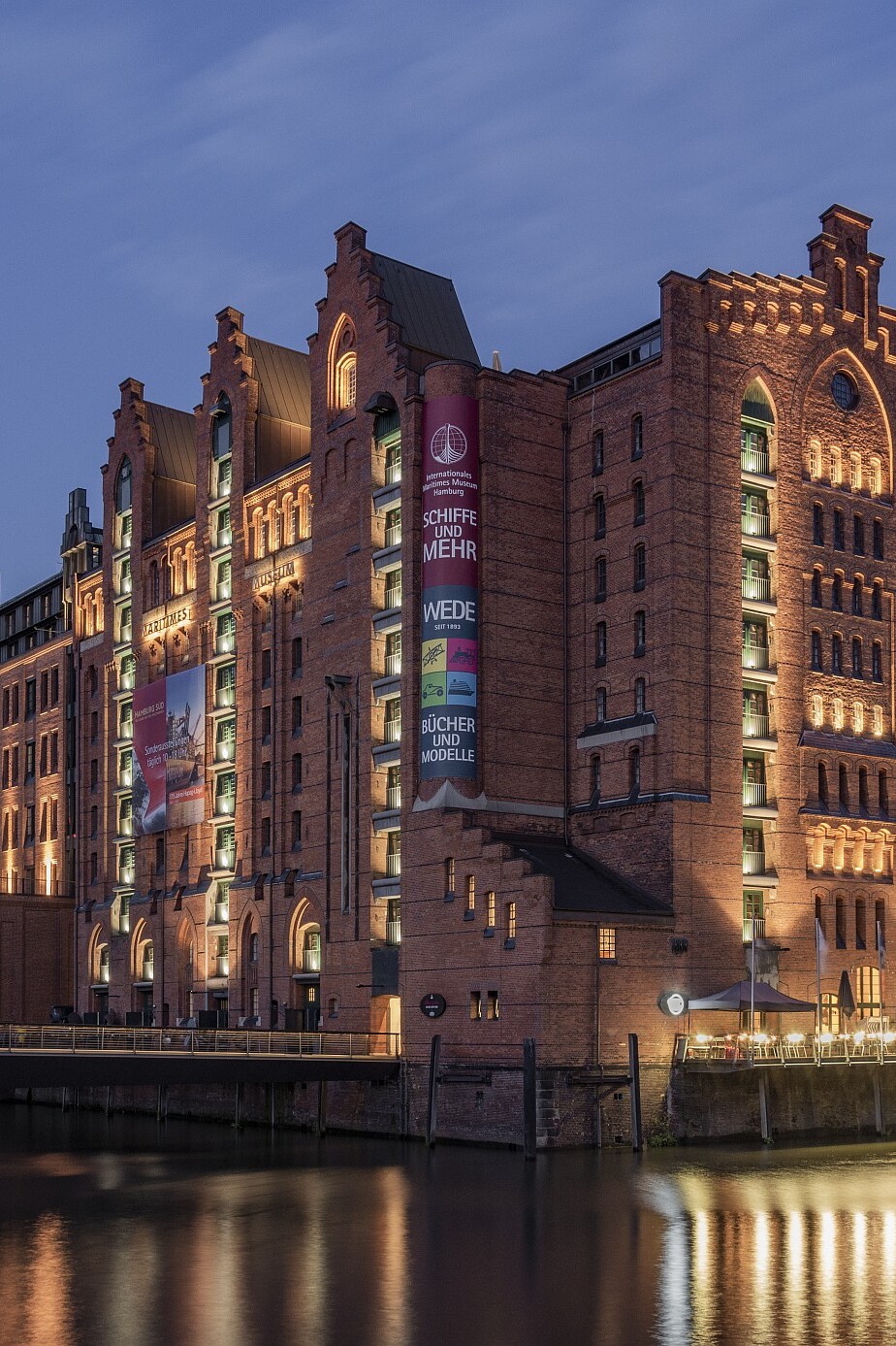Beleuchtetes historisches rotbraunes Backsteingebäude im Lagerhausstil am Wasser, mit Lichterspiegelung auf der Oberfläche