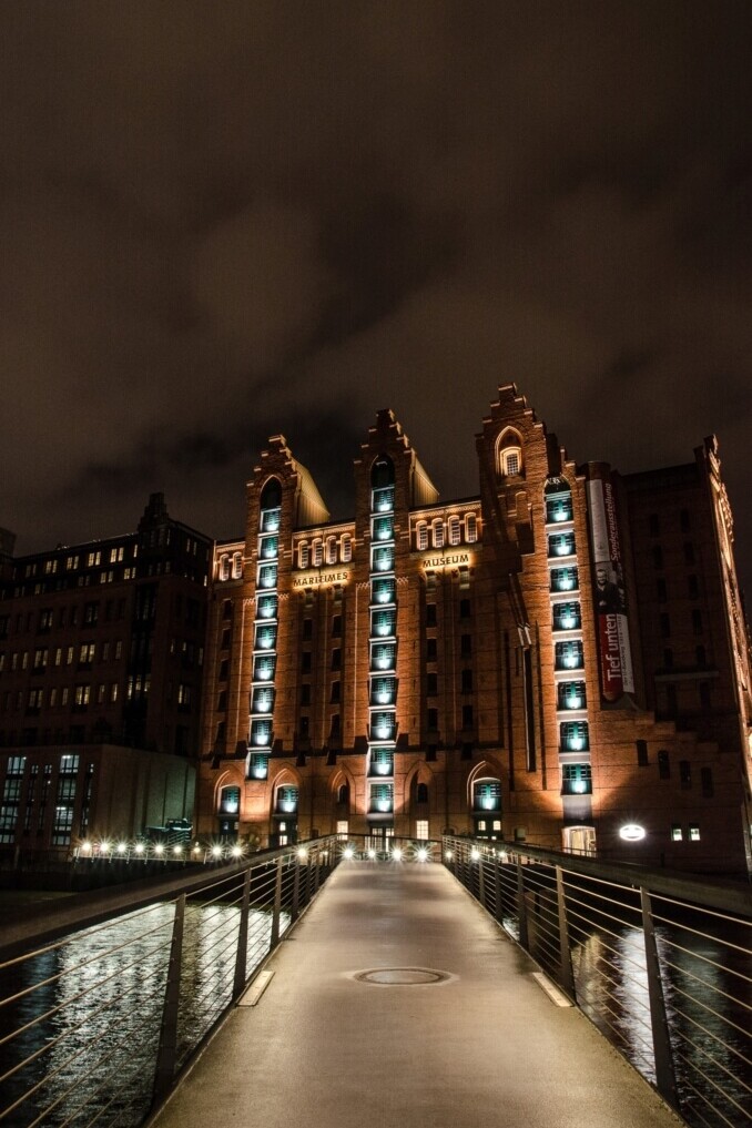 Ein beleuchteter Fußgängersteg führt über einen Kanal zu einem hohen roten Backsteingebäude mit turmartigen Erkern und vertikalen Lichtstreifen bei Nacht.