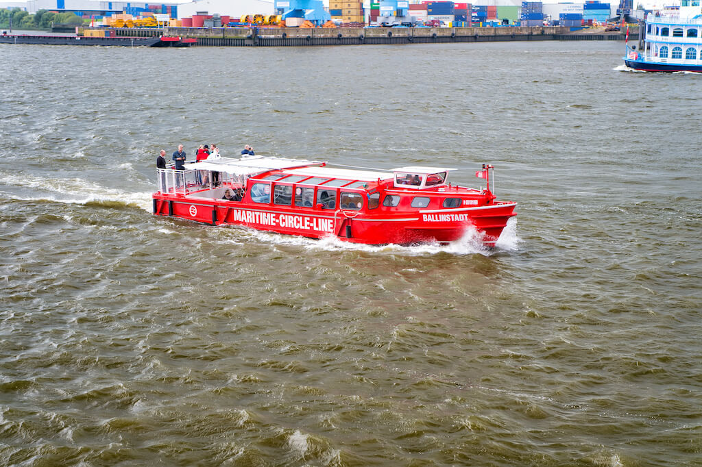 Rotes Ausflugsschiff der Maritime Circle Line mit Passagieren fährt über wellige Hafenwasser vor einem Containerterminal.