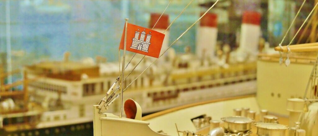 Rote Flagge mit weißer Burg auf dem Deck eines Schiffsmodells, im Hintergrund unscharfe Modellschiffe.