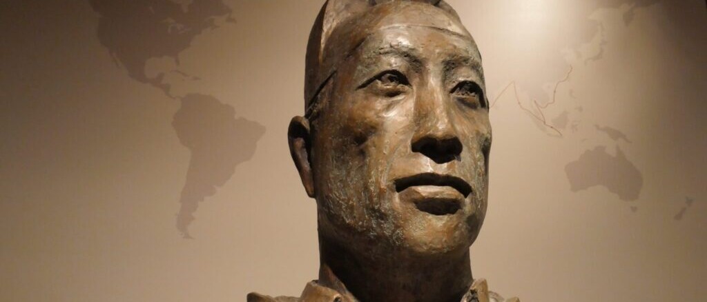 Bronzebüste eines Mannes mit ernstem Blick und Kopfbedeckung, vor einer Wand mit stilisierter Weltkarten-Silhouette
