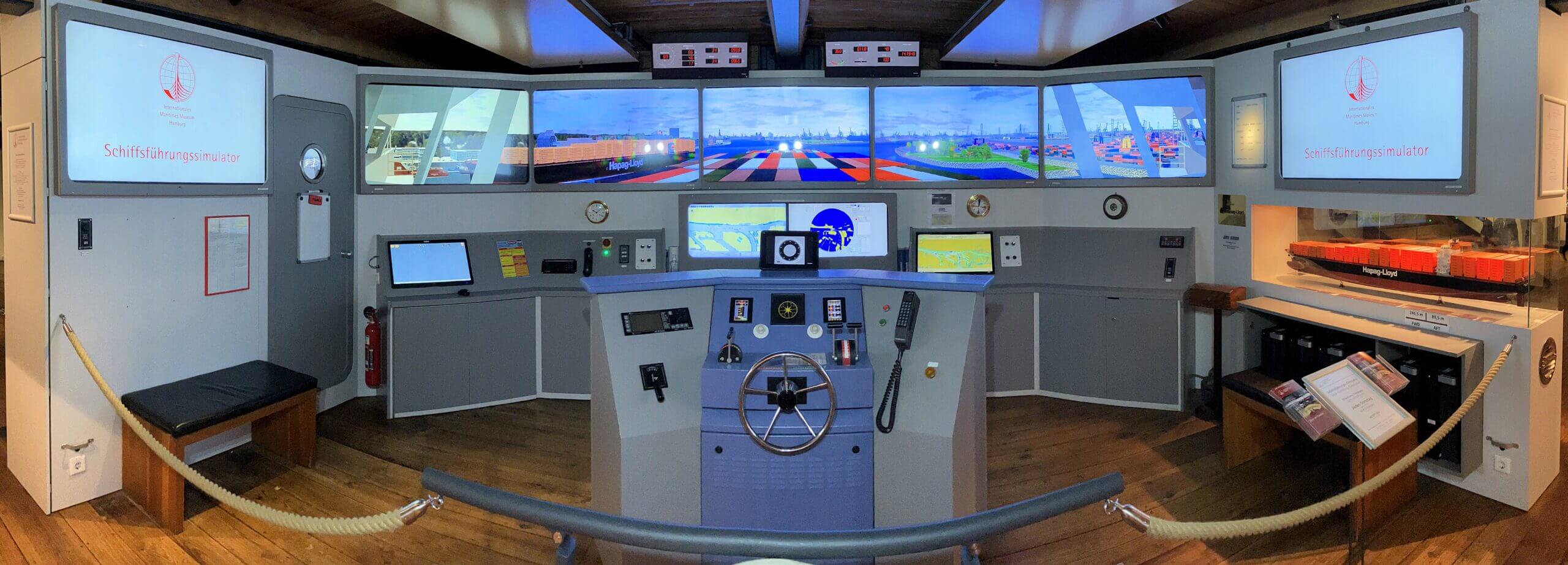 Br&uuml;cken-Navigationssimulator mit Steuerrad, Konsolen und Panoramabildschirmen, rechts ein Modellcontainerschiff.