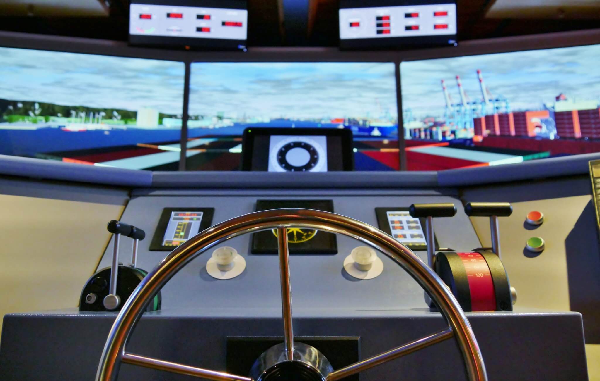 Blick über ein poliertes Metallsteuerrad auf die Brücke eines Schiffs-Simulators mit Schubhebeln, Knöpfen und mehreren Bildschirmen, die einen Hafen mit Containern zeigen.