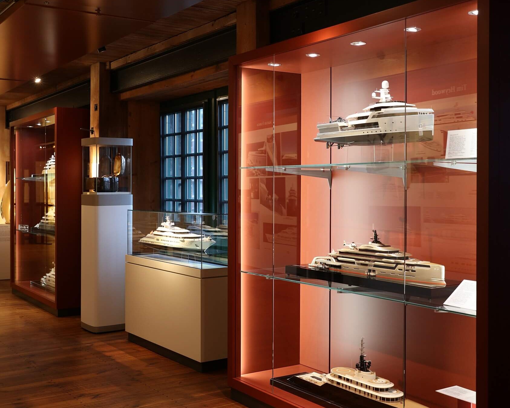 Beleuchteter Ausstellungsraum mit warmem Holzfußboden und -decke, in dem hinter Glasvitrinen mehrere detaillierte Schiffs- und Yachtmodelle vor rot-orangem Hintergrund präsentiert werden.