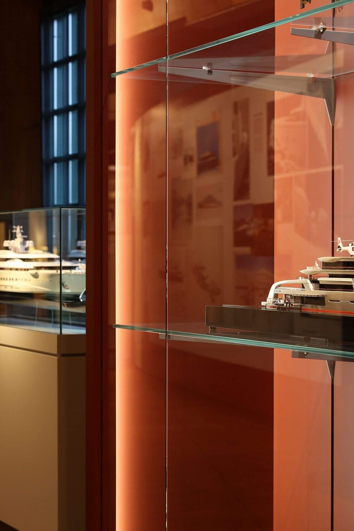 Modell einer modernen Luxus‑Yacht in einer Glasvitrine auf einem Glasregal in einem warm beleuchteten Museumssaal mit roten Wänden, daneben weitere Schiffsmodelle in Vitrinen.