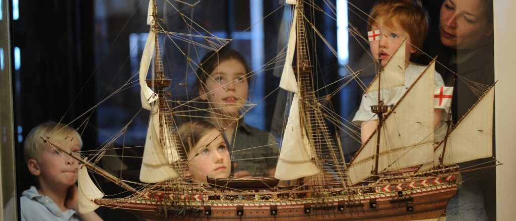 Kinder und Frau betrachten ein detailliertes Segelschiffsmodell mit Takelage und weißen Segeln hinter einer Vitrine.