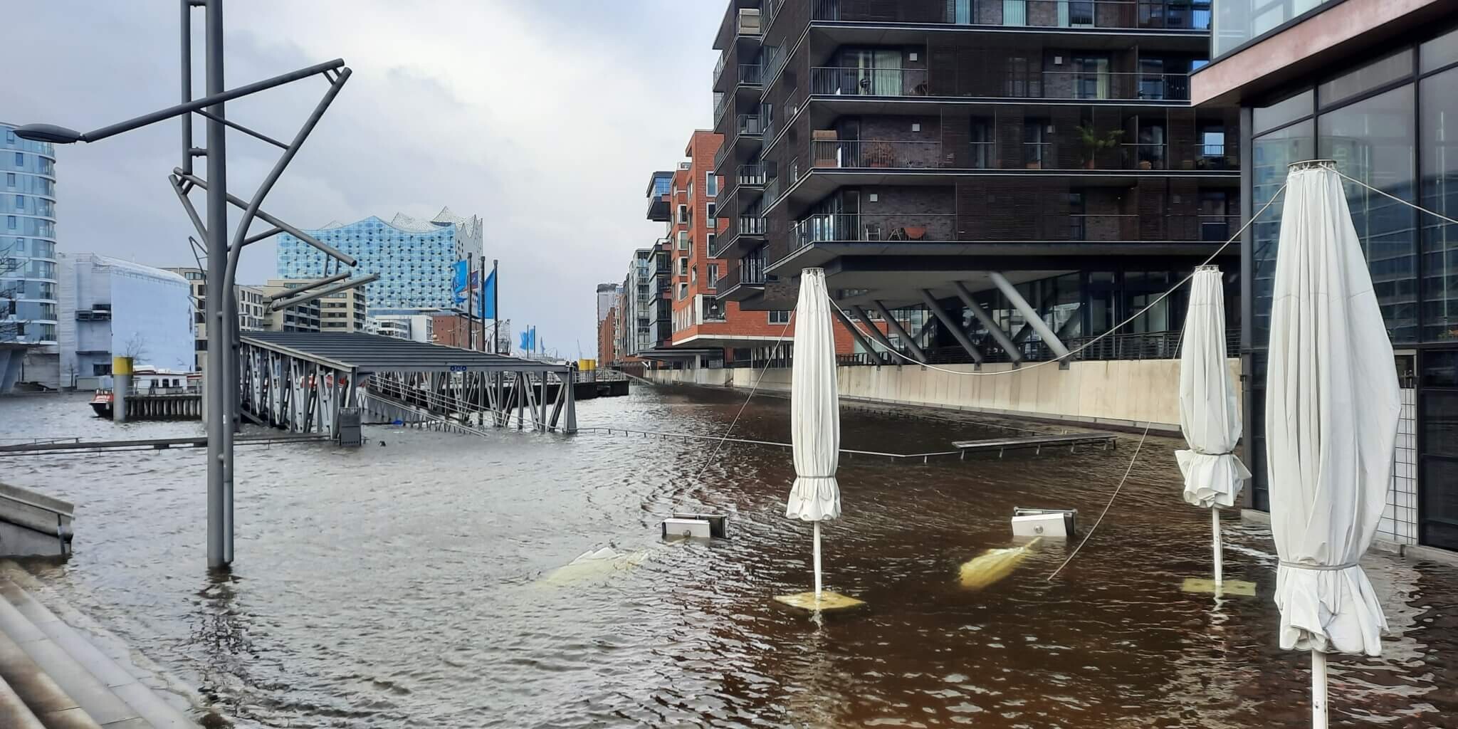 &Uuml;berfluteter Hafen mit teilweise unter Wasser stehenden, gebundenen Sonnenschirmen, moderner Wohnbebauung und der Elbphilharmonie unter grauem Himmel.
