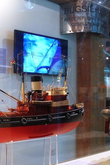 Zwei detaillierte Schlepper-Modelle in einer Museumsvitrine mit roten und schwarzen Rümpfen, am Heck des linken Modells eine deutsche Flagge; im Hintergrund ein maritimes Gemälde, ein blauer Monitor und Spiegelungen auf der Glasoberfläche.