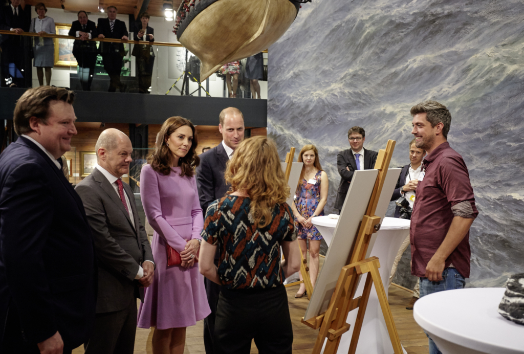 Gruppe gut gekleideter Besucher in einer Ausstellung vor Staffeleien und großem Meeresgemälde; ein Künstler erklärt lächelnd, ein Boot hängt darüber.