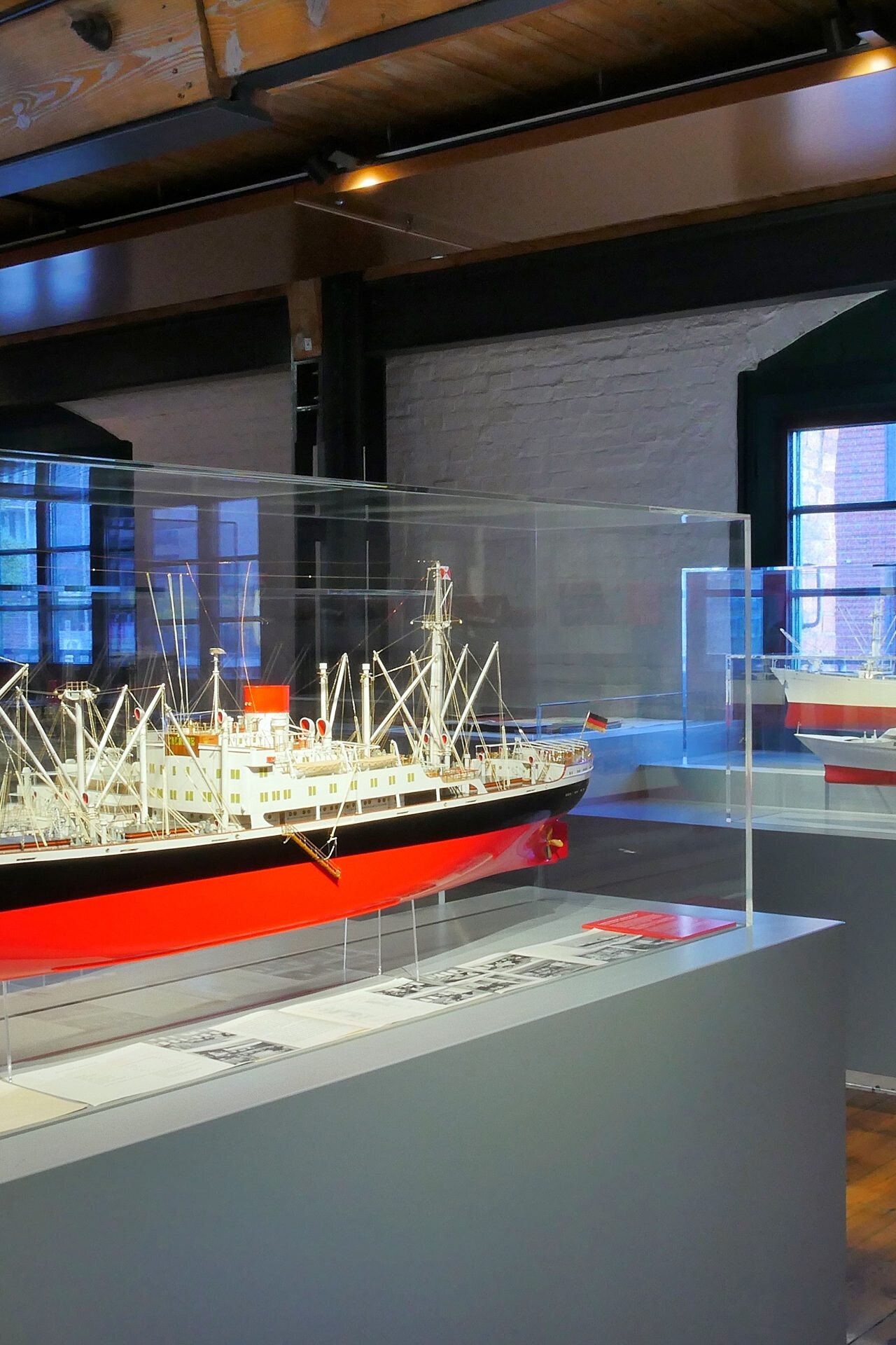 Ausstellungsraum mit gläsernen Vitrinen, in denen detailreiche Schiffsmodelle mit rot‑schwarzen Rümpfen stehen; große Fenster, Holzbalken und Holzfußboden sichtbar.