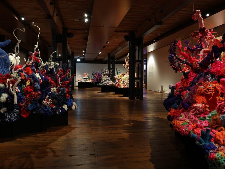 Blick in eine dunkle Galerie mit gro&szlig;formatigen, bunten H&auml;kelinstallationen, die wie Korallenriffe in Rot-, Blau- und Oranget&ouml;nen auf Podesten wachsen.