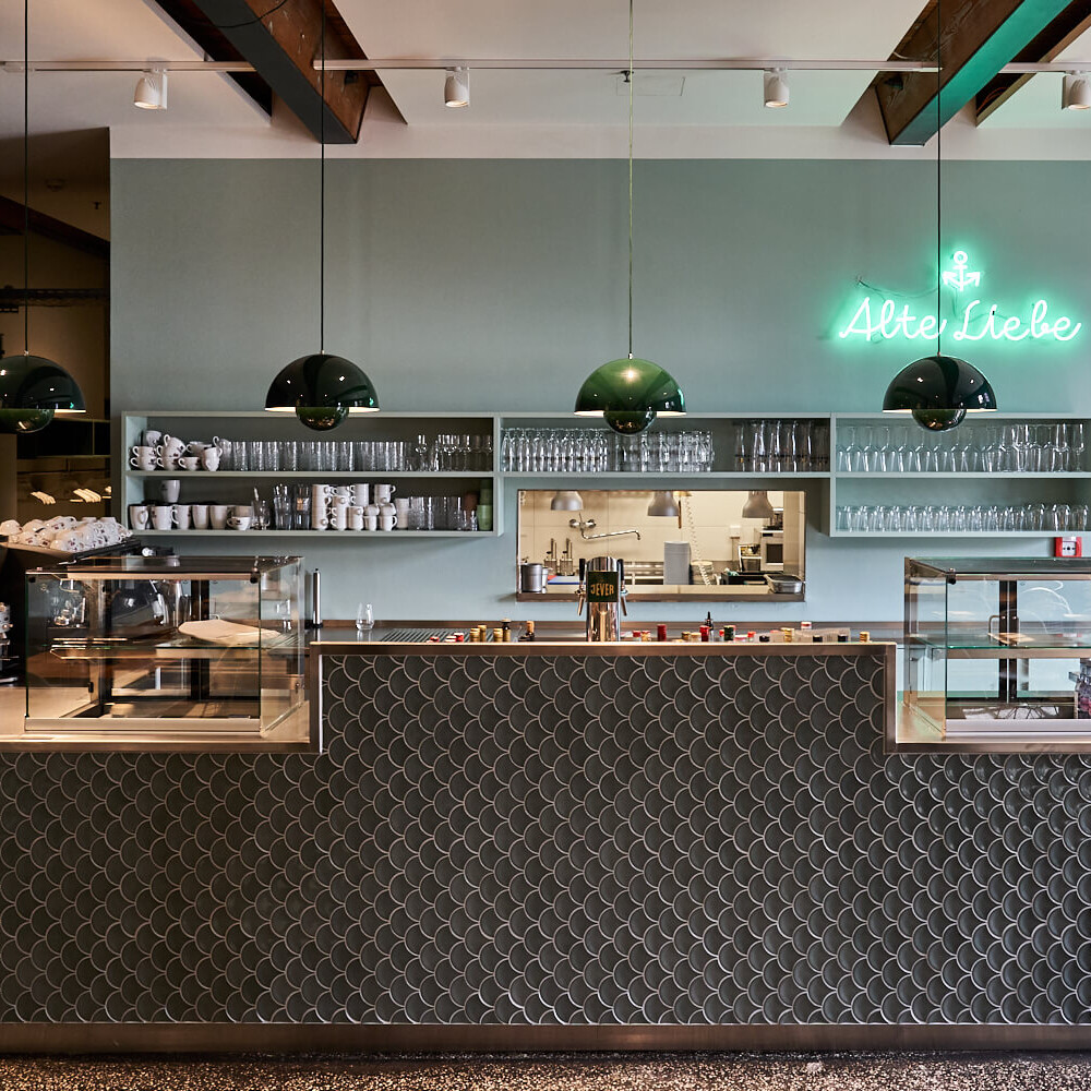 Blick auf die Theke eines Cafés mit geschwungenen Fischschuppenfliesen, zwei Glastheken, grünen Hängelampen, offenen Regalen mit Tassen und Gläsern sowie einem grünen Neon-Schriftzug „Alte Liebe“ über einer Durchreiche zur Küche.