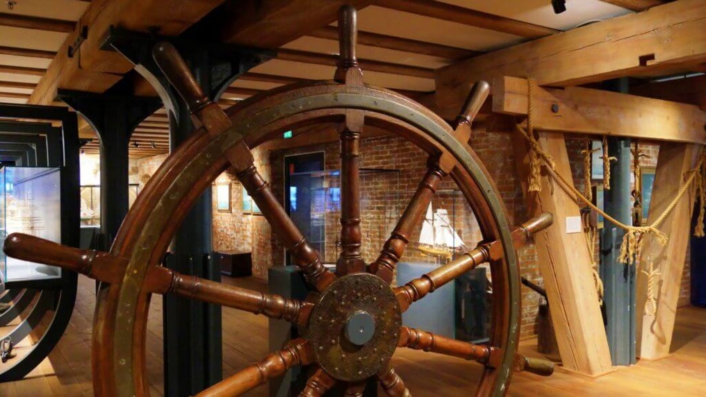 Gro&szlig;es h&ouml;lzernes Schiffssteuerrad im Vordergrund eines Museumsausstellungsraums mit sichtbaren Holztr&auml;gern, Ziegelwand, einem Schiffsmodell im Hintergrund und aufgeh&auml;ngten Tauelementen.